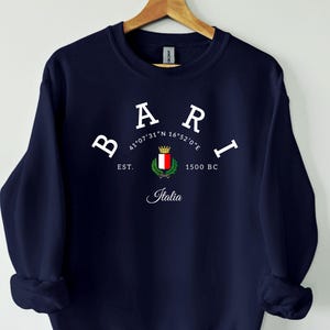 以下が含まれることがあります： 白いアーチ状の文字で「BARI」と書かれたネイビーブルーのスウェットシャツ。その下に「41°07'31"N 16°52'0"E」、「EST. 1500 BC」と「Italia」の文字。スウェットシャツは木製のハンガーに吊るされています。