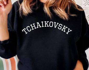 May include: Black sweatshirt with the word "TCHAIKOVSKY" arched across the chest in white lettering. The sweatshirt is a classic crew neck style. The person in the image is wearing gray sweatpants.