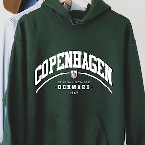May include: Dark green hooded sweatshirt with the word "Copenhagen" in white arched text. Below the text are the words "-DENMARK-" and "1167". The sweatshirt has a front pocket and a hood. A brown hat and a blue shirt hang on the rack behind the sweatshirt.