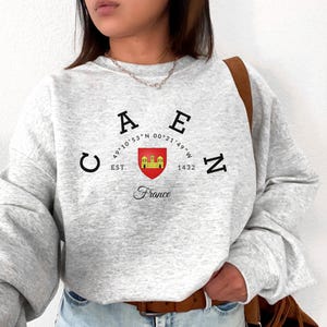 Può includere: Una felpa grigio chiaro con la scritta "CAEN" in lettere nere, uno stemma e la parola "France" in corsivo. La felpa ha maniche lunghe e girocollo.