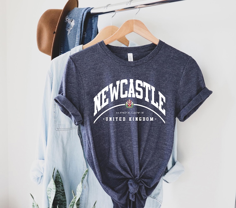 Op de afbeelding: Een marineblauw t-shirt met het woord "NEWCASTLE" in witte gebogen letters. Daaronder een afbeelding en de woorden "UNITED KINGDOM". Het shirt hangt aan een houten hanger met andere kleding.