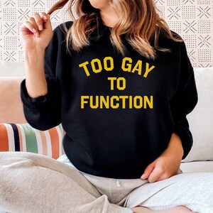 Op de afbeelding: Zwart sweatshirt met de tekst "Too Gay To Function" in gele letters.