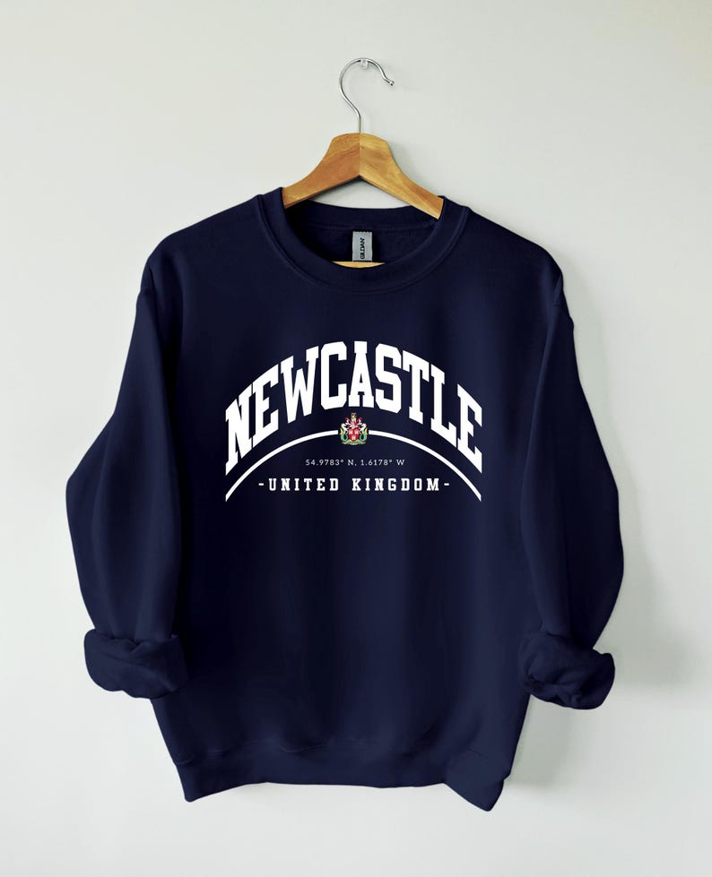 K&ouml;nnte beinhalten: Ein marineblaues Sweatshirt mit dem Wort "NEWCASTLE" in wei&szlig;er, bogenf&ouml;rmiger Schrift. Darunter ein kleines Emblem und der Text "- UNITED KINGDOM -" in Wei&szlig;. Das Sweatshirt h&auml;ngt an einem Holzkleiderb&uuml;gel.