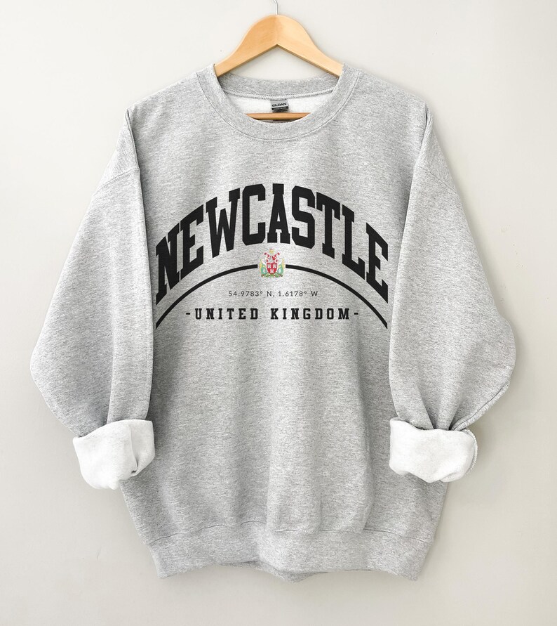 May include: A light gray sweatshirt with the word "NEWCASTLE" arched across the chest in black block letters. Below the text is a crest and the words "UNITED KINGDOM". The sleeves are cuffed, revealing white fabric.