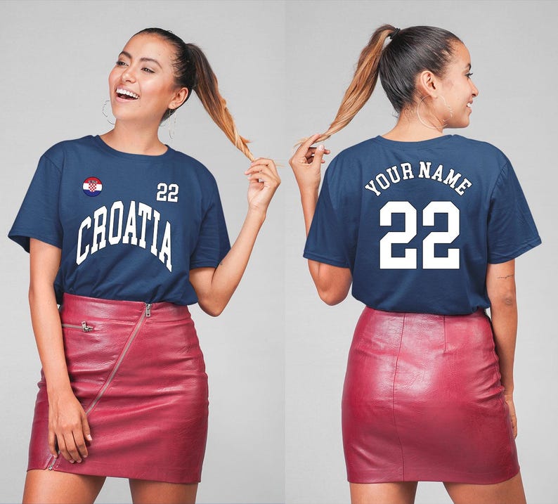 May include: Navy blue t-shirt with "CROATIA" in white arched lettering and the number "22". The Croatian flag is on the left chest. The back of the shirt has "YOUR NAME" and "22" in white. The model is wearing a red skirt.