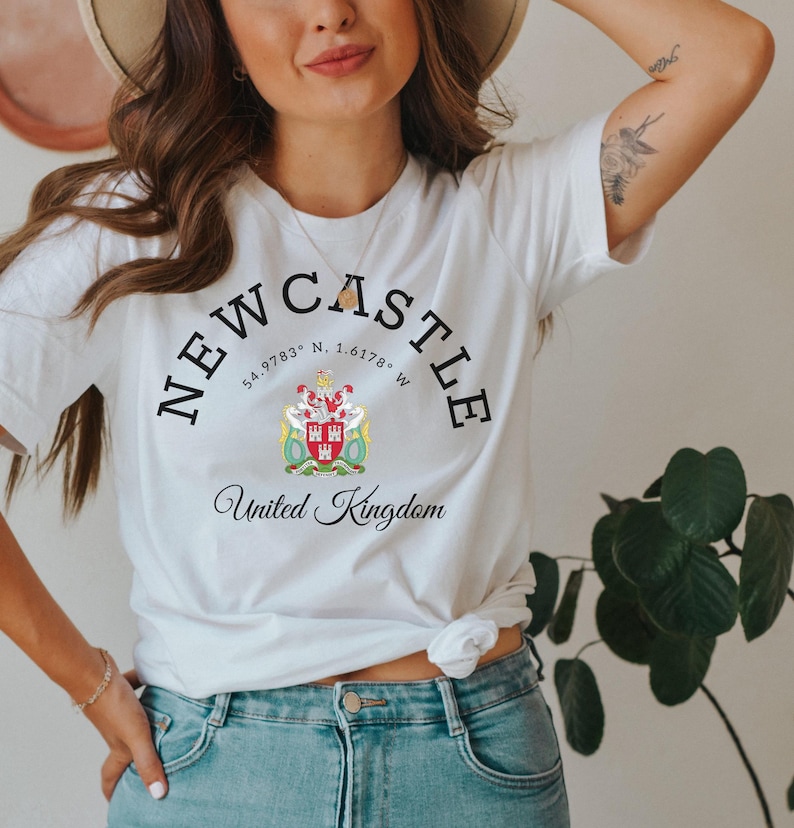 May include: White t-shirt with the word "NEWCASTLE" arched across the top, and "United Kingdom" below. The shirt features a crest with a red and white design. The shirt is worn with blue jeans.