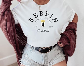 Puede incluir: Camiseta blanca con la palabra "BERLIN" en negro, arqueada sobre un escudo con un oso y una corona. Debajo del escudo están las palabras "EST. 1237" y "Deutschland". La camiseta se usa con pantalones cortos de mezclilla y una rebeca burdeos.