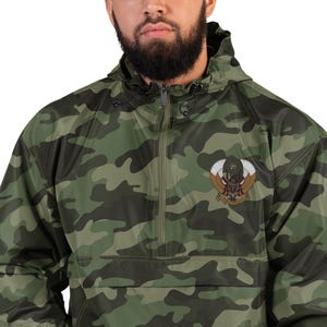 May include: A green and brown camouflage anorak jacket with a hood and a half-zip closure. The jacket features an embroidered emblem on the chest with a central figure, wings, and a banner. The jacket is made of a lightweight material.