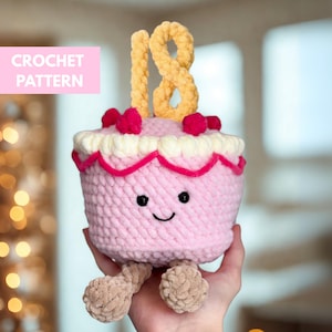 Op de afbeelding: Een roze gehaakte taart met een lachend gezicht, bekroond met het gele nummer "18" en rode besachtige versieringen. De taart heeft een witte en roze rand en kleine bruine pootjes. De tekst "CROCHET PATTERN" staat in een roze rechthoek.