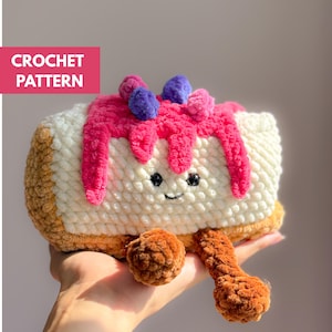 Puede incluir: Tarta de ganchillo hecha a mano con una cara sonriente, glaseado rosa y acentos de bayas moradas y rosas. La tarta tiene secciones marrones y blancas y dos patas marrones. El texto "CROCHET PATTERN" está en un rectángulo rosa.