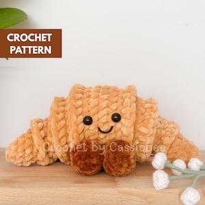 以下が含まれることがあります： ベージュと茶色の色合いの、かぎ針編みのクロワッサンのおもちゃ。おもちゃには、黒いボタンの目と縫い付けられた口を持つ笑顔の顔があります。「CROCHET PATTERN」という文字が左上の茶色のボックスにあります。「Crochet by Cassiopee」という文字がおもちゃにあります。
