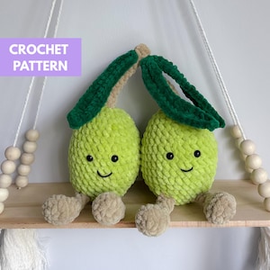 Könnte beinhalten: Zwei gehäkelte Fruchtspielzeuge in Limettengrün mit lächelnden Gesichtern, grünen Blättern und beigefarbenen Füßen, die auf einem Holzregal sitzen. Der Text "CROCHET PATTERN" befindet sich in einem lila Rechteck. Das Regal ist an einem weißen Seil aufgehängt.