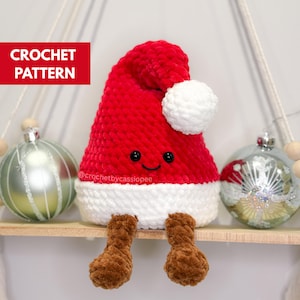 May include: A red and white crocheted Santa hat with a smiling face and brown legs. The hat has a white pom-pom and trim. The text "CROCHET PATTERN" is in a red rectangle. The hat is sitting on a wooden shelf with Christmas ornaments.