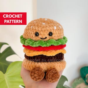 Pode incluir: Um brinquedo de hambúrguer de crochê com um pão bege com sementes de gergelim brancas bordadas, uma camada de alface verde, uma fatia de tomate vermelho, uma camada de queijo laranja, um hambúrguer marrom escuro e dois pés marrons. Texto CROCHET PATTERN.