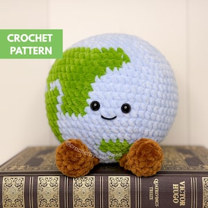 Op de afbeelding: Een gehaakte aarde-knuffel met een lachend gezicht, blauwe oceanen en groene continenten. De knuffel heeft bruine voetjes en is voorzien van het label "CROCHET PATTERN". De aarde zit op een boek.