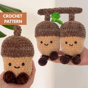 May include: Two crochet acorn plushies made from soft brown and tan yarn. One is a single acorn, and the other is a pair attached to a branch with a green leaf. Both have black safety eyes and stitched smiles. Text reads CROCHET PATTERN.