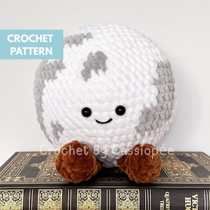 Peut inclure: Une lune en peluche crochetée avec des taches grises, des yeux noirs et une bouche souriante. La lune a des pieds marron et est posée sur un livre. Le texte "CROCHET PATTERN" est dans un encadré bleu. Le texte "Crochet By Cass" est en dessous.