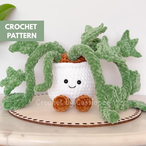 Op de afbeelding: Een gehaakte plant in een witte pot met een lachend gezicht en bruine voetjes. De plant heeft groene, fuzzy bladeren en staat op een houten basis. De tekst "CROCHET PATTERN" staat in een groen vak linksboven. De tekst "Crochet by Cassiopee" staat rechtsonder.
