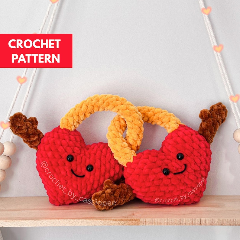 May include: Two red crochet hearts with smiling faces and brown antlers are connected by a yellow pretzel-shaped piece. The text "CROCHET PATTERN" is in a red box in the upper left corner. The hearts are on a wooden shelf.