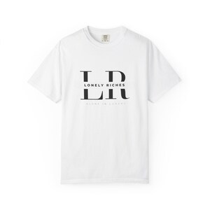 May include: White t-shirt with a crew neck and short sleeves. The shirt displays the text "LONELY RICHES" in a black, stylized font. Beneath this is the phrase "ALONE IN LUXURY" in a smaller font.