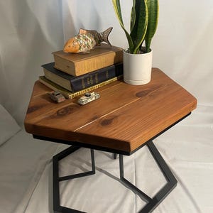 May include: A hexagonal wooden table with a black metal base. The table top is made of natural wood with a rustic finish. A potted snake plant, a stack of books, and a decorative fish figurine are on the table.