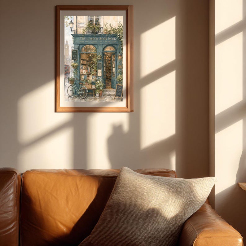 A framed print of "The London Book Nook" hangs on a light-colored wall above a brown leather sofa with a patterned pillow, with sunlight casting shadows.
