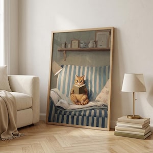 May include: A framed print shows an orange tabby cat reading a book on a blue and white striped sofa. The artwork is in a wooden frame and features a shelf with decorative items. A lamp and books are in the foreground.