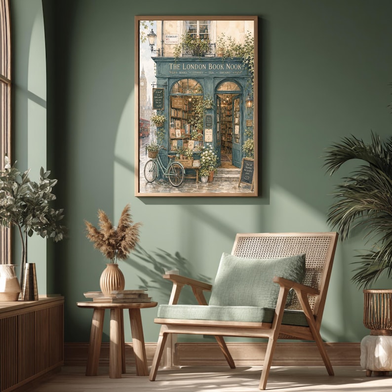 A framed print of a watercolor illustration of a blue-green storefront with "THE LONDON BOOK NOOK" above the door, a bicycle parked outside, and Big Ben in the background, hanging on a dark green wall above a wooden chair and side table.