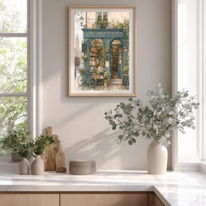 A framed print of "The London Book Nook" hangs on a white wall in a kitchen, with plants on the counter and a window to the left.