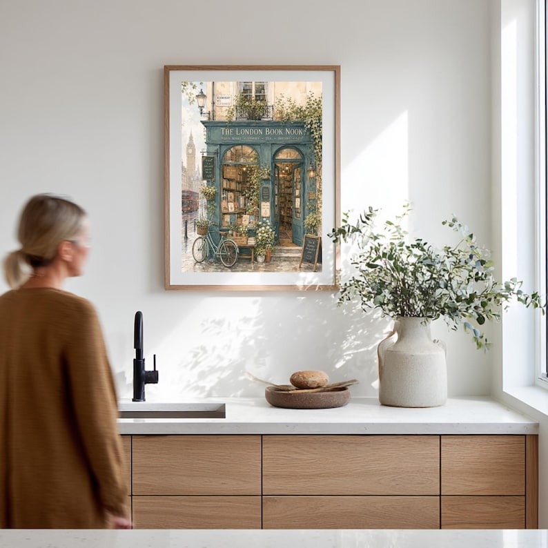 A framed print of "The London Book Nook" hangs on a white wall above a kitchen counter, with a person in a brown sweater walking away.