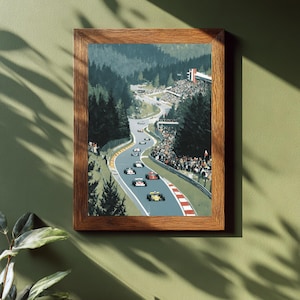 Könnte beinhalten: Ein gerahmter Druck einer Oldtimer-Rennwagenszene. Das Kunstwerk zeigt Rennwagen auf einer kurvenreichen Strecke durch einen grünen Wald. Die Strecke hat rot-weiß gestreifte Bordüren. Der Rahmen ist aus braunem Holz.