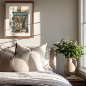 A framed print of a watercolor illustration of a blue-green storefront with "THE LONDON BOOK NOOK" above the door, a bicycle parked outside, and Big Ben in the background, hanging on a light gray wall above a bed with beige pillows.