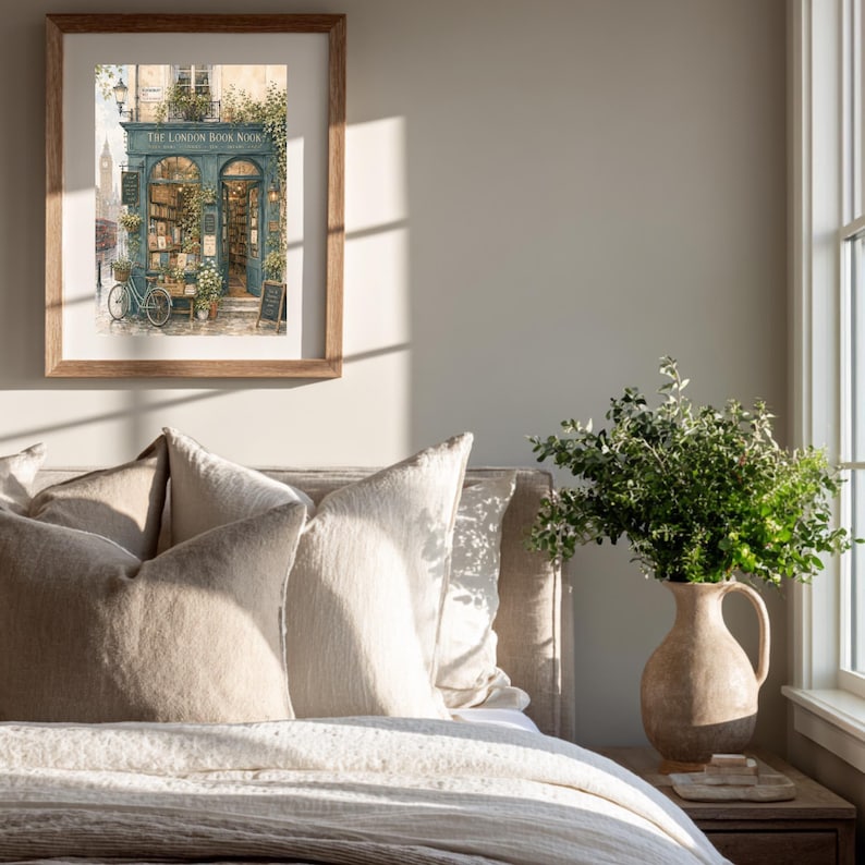 A framed print of a watercolor illustration of a blue-green storefront with "THE LONDON BOOK NOOK" above the door, a bicycle parked outside, and Big Ben in the background, hanging on a light gray wall above a bed with beige pillows.