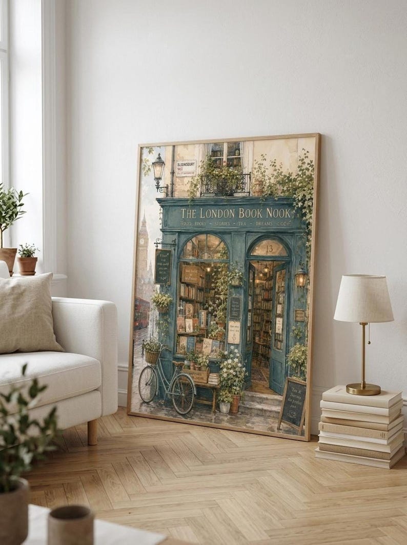 A framed print of a watercolor illustration of a blue-green storefront with "THE LONDON BOOK NOOK" above the door, a bicycle parked outside, and Big Ben in the background, leaning against a white sofa in a room with light wood floors.