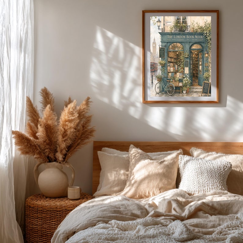 A framed print of "The London Book Nook" hangs on a white wall above a bed with white linens, with a vase of dried grasses on a side table.