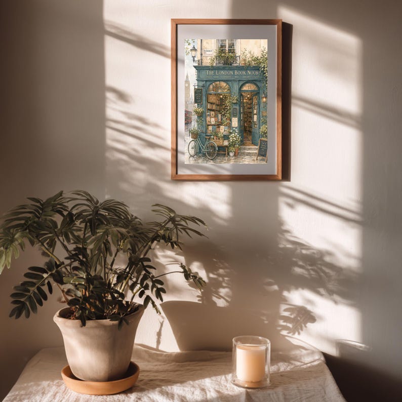 A framed print of "The London Book Nook" hangs on a wall with sunlight and shadows, above a table with a potted plant and a candle.