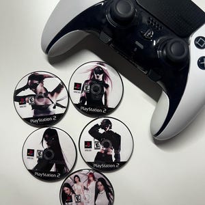 Puede incluir: Un mando blanco y negro de PlayStation 5 descansa cerca de cinco posavasos circulares. Cada posavasos presenta a una mujer con una estética oscura, el logotipo de PlayStation 2 y la calificación "E". Los posavasos tienen un fondo blanco.