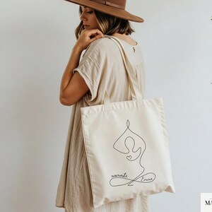 May include: Cream-colored tote bag featuring a black line drawing of a yoga pose with a heart. The words "namaste Emma" are written in cursive below the figure. The bag has two straps and is held by a person.