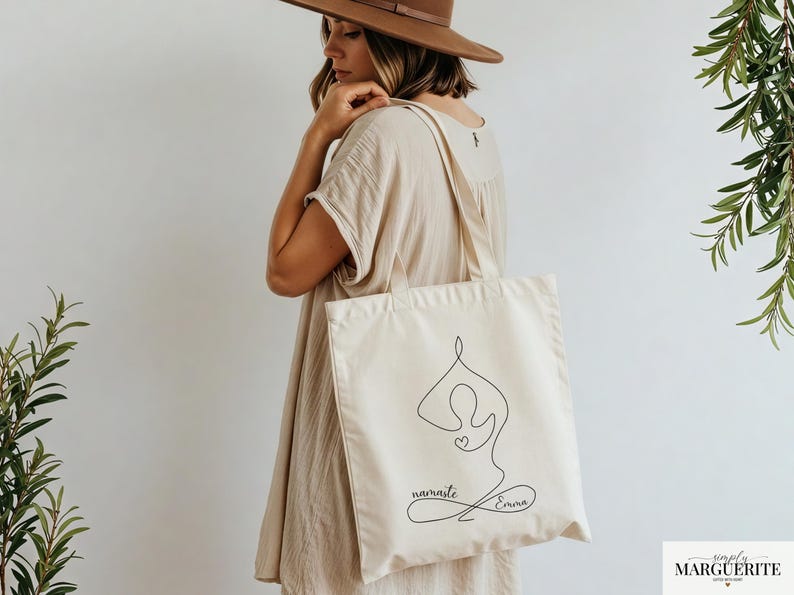 May include: Cream-colored tote bag featuring a black line drawing of a yoga pose with a heart. The words "namaste Emma" are written in cursive below the figure. The bag has two straps and is held by a person.