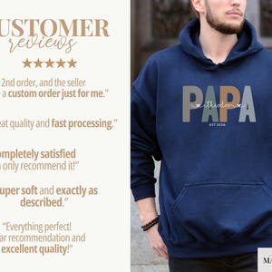 Puede incluir: Sudadera con capucha azul marino con la palabra "PAPA" en letras grises, naranjas y azules. El nombre "theodor" est&aacute; escrito en blanco, con "EST 2026" debajo. La sudadera tiene un bolsillo delantero y una capucha con cord&oacute;n.