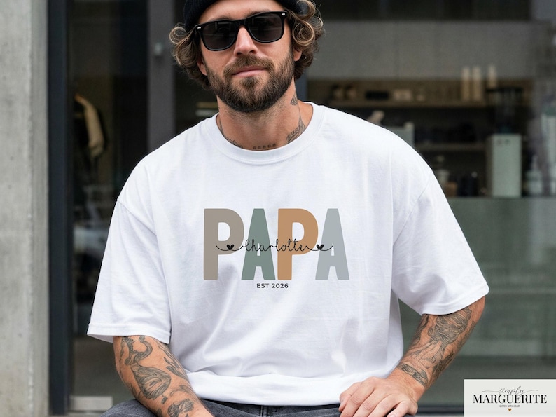 Puede incluir: Camiseta blanca con la palabra "PAPA" en letras de bloque marrones, verdes y grises. Debajo de la palabra "PAPA" est&aacute; el nombre "Charlotte" en cursiva, con dos peque&ntilde;os corazones. El texto "EST 2026" est&aacute; debajo del nombre.