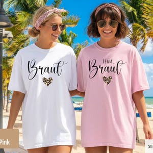 Pu&ograve; includere: Due donne indossano magliette bianche e rosa chiaro. La maglietta bianca dice "Braut" e quella rosa "Team Braut". Entrambe hanno un carattere script e un design a cuore leopardato.