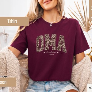 May include: Maroon T-shirt with the word "OMA" in a leopard print design. Below the word "OMA" is the name "charlotte" with two hearts and the text "EST 2026". The shirt is made of 100% cotton.
