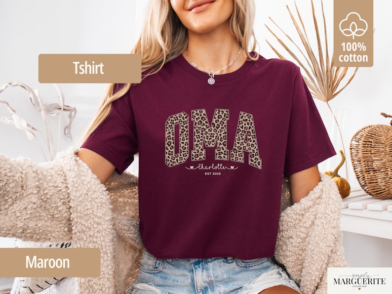 May include: Maroon T-shirt with the word "OMA" in a leopard print design. Below the word "OMA" is the name "charlotte" with two hearts and the text "EST 2026". The shirt is made of 100% cotton.
