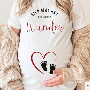 May include: White maternity t-shirt with the German text "Hier wächst ein kleines Wunder" in black and red script. A red heart with black footprints is printed on the front. The shirt is worn with a beige cardigan.