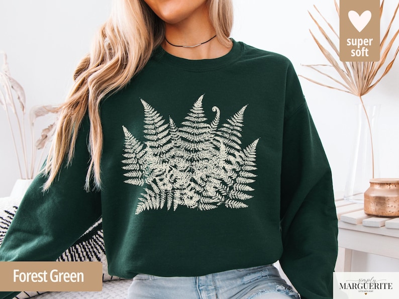 Puede incluir: Sudadera verde bosque con un dise&ntilde;o de helecho crema. La ilustraci&oacute;n bot&aacute;nica est&aacute; centrada en el pecho. La sudadera tiene cuello redondo y mangas largas. Las palabras "super soft" est&aacute;n en un coraz&oacute;n en la esquina superior derecha.
