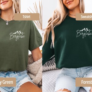 May include: Military green t-shirt and forest green sweatshirt with the word "Bergliebe" and a mountain graphic in white. The t-shirt is short-sleeved, and the sweatshirt is long-sleeved. Both are casual wear.