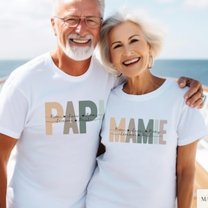 May include: A smiling couple wearing matching white t-shirts with pastel text. One shirt says "PAPI" and the other says "MAMIE". Each shirt has names below the text. The background shows a blue ocean and a white boat railing.