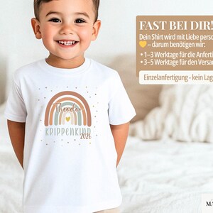 May include: A white short-sleeved t-shirt featuring a graphic of a multi-colored rainbow in shades of brown, blue, and cream. The design includes the name Theodor, the text KRIPPENKIND, the year 2026, a small yellow heart, and scattered gold dots.