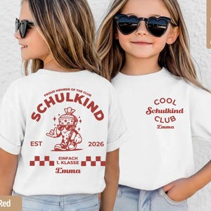May include: A white t-shirt featuring red graphic prints. The back displays a cartoon character in a cone, the text "PROUD MEMBER OF THE CLUB SCHULKIND EST 2026 EINFACH 1. KLASSE" and the name "Emma". The front features "COOL Schulkind CLUB Emma".